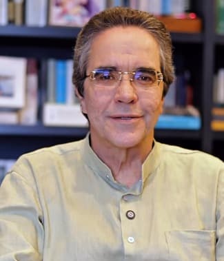 Maurício Rands em retrato, de camisa clara e óculos, em ambiente de estante de livros